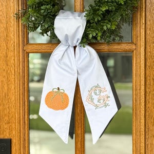 May include: A white fabric door hanger featuring two long, pointed tails. The left tail displays an embroidered orange pumpkin with a green stem and polka dot pattern, while the right tail features an embroidered ornate letter G in peach and green.