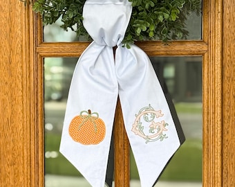 May include: A white fabric door hanger featuring two long, pointed tails. The left tail displays an embroidered orange pumpkin with white polka dots and a green stem, while the right tail features an embroidered ornate letter G in peach and green.