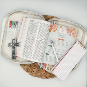 May include: An open Bible with colorful page markers and a study guide, resting inside a floral-patterned case. A small notepad, pens, and a cross-shaped embellishment are also visible, suggesting a devotional or study set.