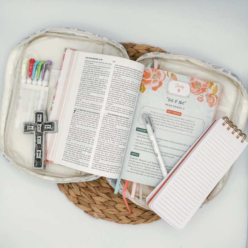 May include: An open Bible with colorful page markers and a study guide, resting inside a floral-patterned case. A small notepad, pens, and a cross-shaped embellishment are also visible, suggesting a devotional or study set.