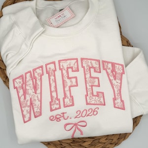 May include: A cream-colored crewneck sweatshirt featuring the embroidered text WIFEY in large block letters with a pink floral pattern fill. Below the text, the words est. 2026 and a small pink bow are embroidered in a matching pink thread.