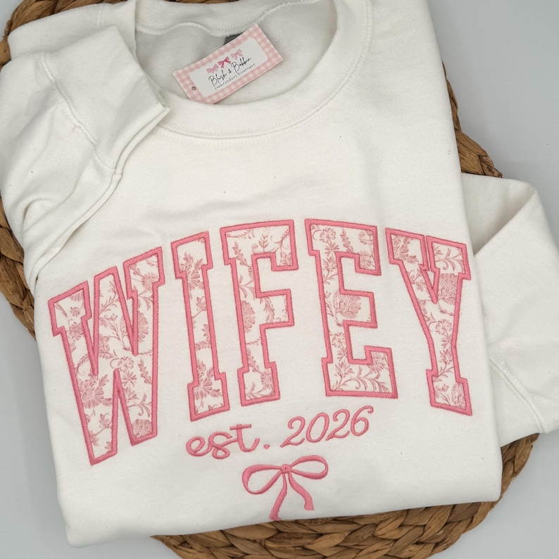 May include: A cream-colored crewneck sweatshirt featuring the embroidered text WIFEY in large block letters with a pink floral pattern fill. Below the text, the words est. 2026 and a small pink bow are embroidered in a matching pink thread.