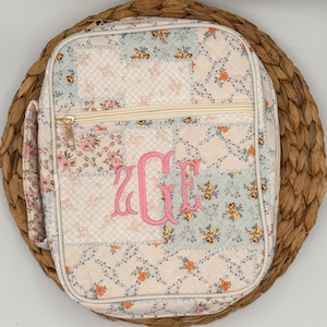 May include: A quilted, floral-patterned lunch bag with a pink monogram. The bag features a patchwork design with various floral prints in pastel colors. It has a zippered closure and a side pocket. The bag is placed on a woven, circular placemat.
