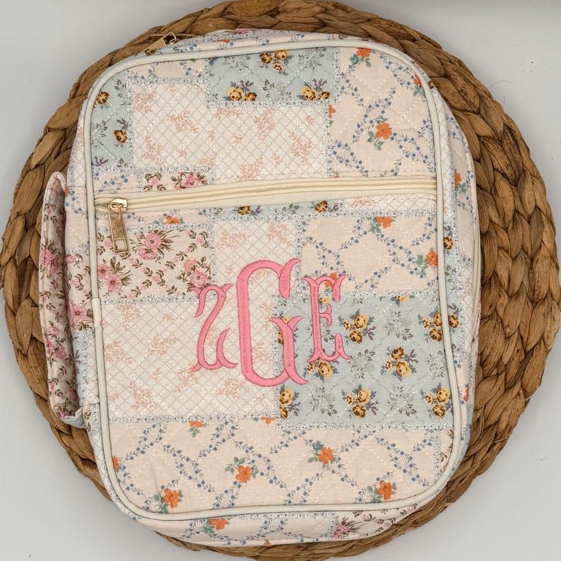 May include: A quilted, floral-patterned lunch bag with a pink monogram. The bag features a patchwork design with various floral prints in pastel colors. It has a zippered closure and a side pocket. The bag is placed on a woven, circular placemat.
