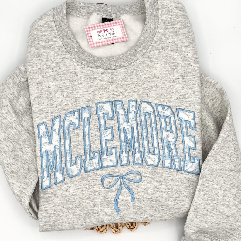 May include: A heather gray crewneck sweatshirt featuring the text MCLEMORE in large, arched, light blue block letters with a white floral pattern inside. Below the text is a light blue embroidered bow. A tag reads Blush & Bobbin.