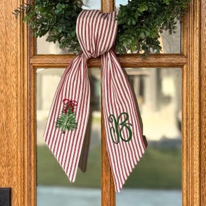 May include: Red and white striped fabric door hanger with a Christmas tree and a green monogram. The fabric is tied in a knot and hangs down. The Christmas tree is green with a red bow. The monogram is green.