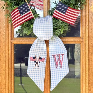 May include: A patriotic wreath with American flags and a blue and white gingham bow. The bow features an embroidered red letter "W" and small American flags. The wreath is displayed on a wooden door.