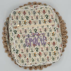 May include: A floral patterned bag with a zipper closure and the name "Gracie" embroidered in purple. The bag has a white trim and is set on a woven placemat. The floral pattern includes red, green, and yellow flowers.