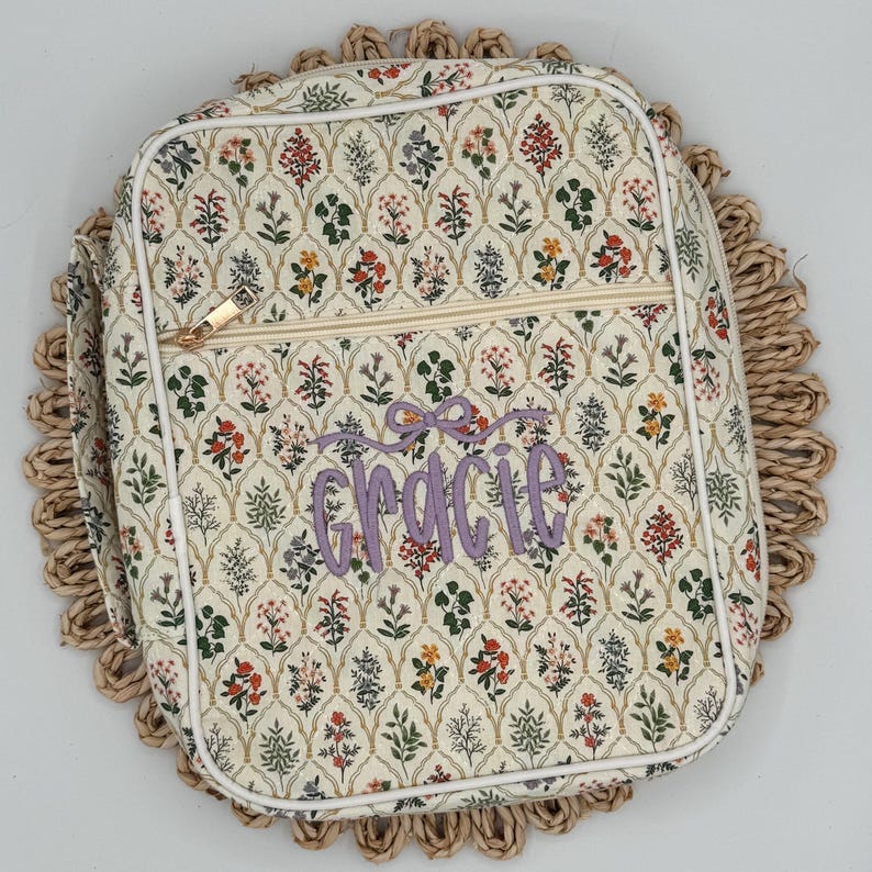 May include: A floral patterned bag with a zipper closure and the name "Gracie" embroidered in purple. The bag has a white trim and is set on a woven placemat. The floral pattern includes red, green, and yellow flowers.