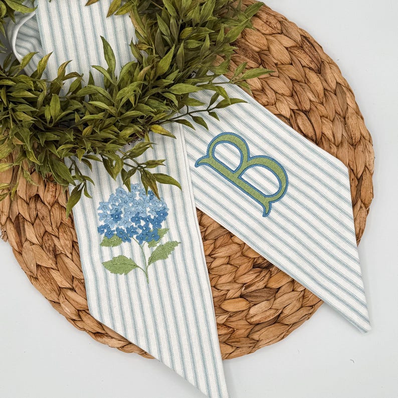 May include: Two decorative fabric pieces with blue and white stripes. One features a blue hydrangea embroidery, the other a green letter "B". The items are arranged on a woven, circular placemat, with green foliage.