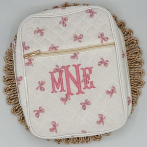 May include: White quilted bag with pink bow accents and a gold zipper. The bag features a pink monogram of the letters "MNE". The bag is sitting on a woven placemat. The bag is rectangular with rounded corners.
