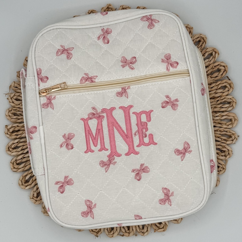 May include: White quilted bag with pink bow accents and a gold zipper. The bag features a pink monogram of the letters "MNE". The bag is sitting on a woven placemat. The bag is rectangular with rounded corners.