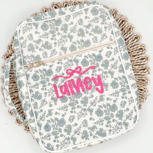 May include: A floral-patterned bag with a white background and blue flowers. The bag has a zipper closure and a handle. The name "Lainey" is embroidered in pink script with a bow above it. The bag is on a woven placemat.