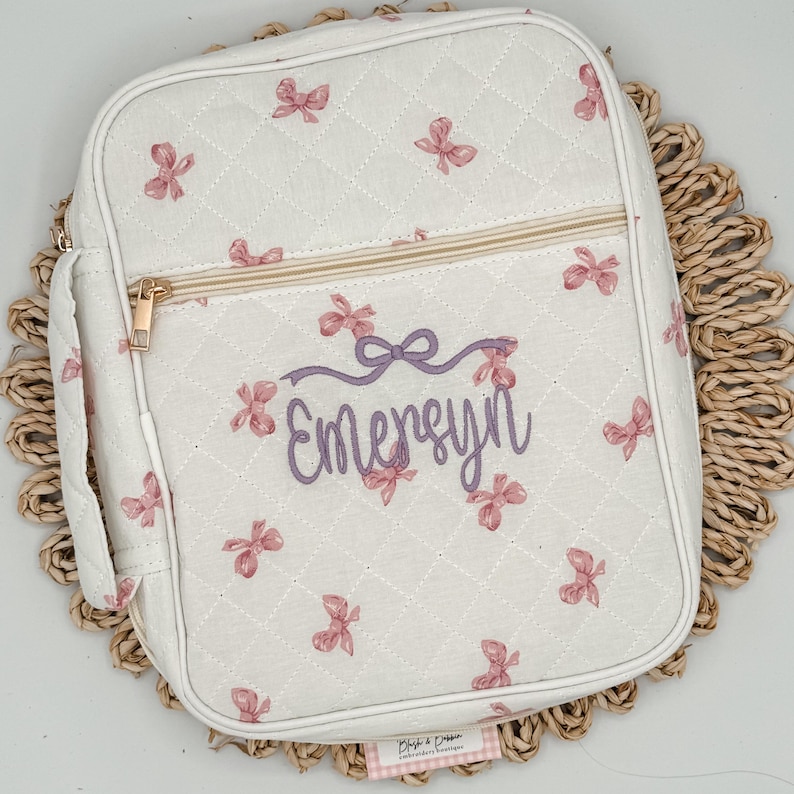 May include: A white quilted lunch bag with a repeating pattern of pink bows. The bag features a gold zipper and a handle. The name "Emersyn" is embroidered in purple script on the front.