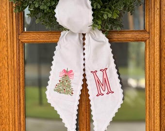 May include: A white fabric door hanger featuring a scalloped edge and a green wreath at the top. The left tail displays an embroidered Christmas tree with a pink bow, while the right tail features a large, embroidered pink letter M in a serif font.