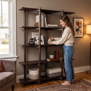 Könnte beinhalten: Ein hohes, dunkelbraunes Holzregal mit mehreren offenen Fächern. Die Regale sind mit verschiedenen Gegenständen bestückt, darunter Bücher, eine geometrische Drahtskulptur, eine Vintage-Kamera, eine kleine Topfpflanze und ein weißer geflochtener Korb.