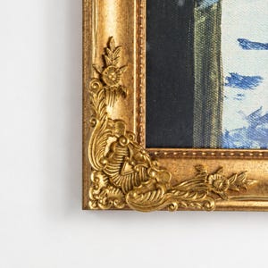May include: A gold-colored picture frame featuring an ornate corner detail with raised floral and scrollwork patterns. The frame has a textured, metallic finish and surrounds a canvas with visible blue and black paint strokes on a white background.