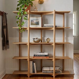 May include: A light brown wooden bookshelf with five tiers featuring vertical support beams. The shelves hold a potted green plant, a framed landscape print, books titled Designing Home, US Modernism, American Craft, The New York School, and Mid-Century Modern Design, a bowl, a clock, and a folded plaid blanket.
