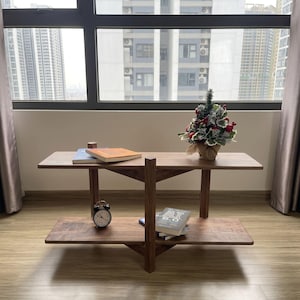 May include: A two-tier wooden console table with a dark brown finish, featuring a central vertical support beam. The top shelf holds a small decorative Christmas tree and two books, while the bottom shelf holds a small analog clock and a book.