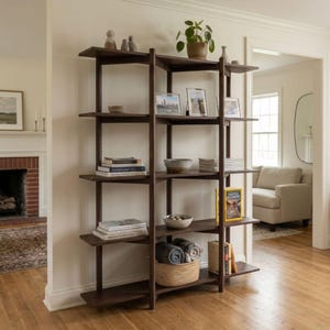 May include: A tall, dark brown wooden bookshelf with five horizontal shelves featuring an open, vertical support structure. The shelves hold various items including books, a potted plant, framed photos, bowls, and a woven basket with textiles.