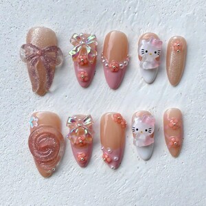 Può includere: Un set di dieci unghie finte con vari design. Alcune unghie presentano una base nude con dettagli rosa, fiocchi e decorazioni floreali. Altre mostrano un design di Hello Kitty e glitter.