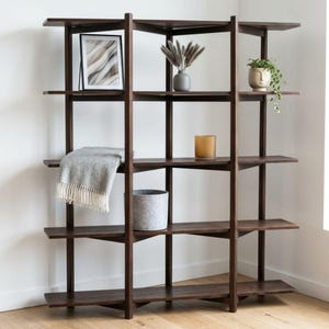 May include: A dark brown wooden bookshelf with five tiers of shelving. The shelves hold a framed abstract print, a grey vase with dried grasses, a small face-shaped planter with a trailing green plant, a grey throw blanket, and a glass candle.