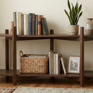 May include: A dark wood two-tier console table holding a row of books, a potted snake plant, and a ceramic vase on the top shelf, with a woven seagrass basket, a second row of books, and a framed photograph on the bottom shelf.