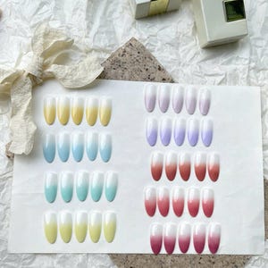 May include: A collection of press-on nails in various ombre color schemes. The colors include yellow, blue, teal, purple, and red, transitioning to white at the tips. The nails are arranged on a white surface, with a neutral background.