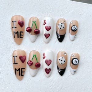 May include: A set of ten artificial nails with various designs. Some nails feature the text "I ❤️ ME" with a heart, cherries, the number 3, dice, and a number 5. The nails are painted in nude, white, and black.