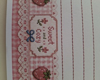 Könnte beinhalten: Ein weißes Briefpapier mit einem rosa karierten Rand mit Spitzenbesatz, roten Erdbeerillustrationen und einem zentralen rechteckigen Rahmen mit dem gestickten Text Sweet and Cozy, akzentuiert durch eine kleine blaue Schleife.