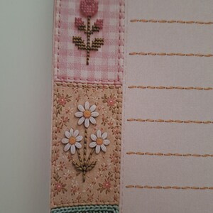 May include: A sheet of stationery featuring a vertical border of patchwork-style squares with floral and crochet patterns in pink, beige, and green. The right side of the paper is light pink with horizontal lines marked by orange dashed stitches.