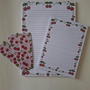 Può includere: Due fogli di carta da lettere bianca a righe e tre segnalibri rettangolari. La carta da lettere presenta un bordo di ciliegie rosse con foglie e viticci verdi. I segnalibri hanno uno sfondo a quadretti rosa e bianco con motivi a ciliegia.