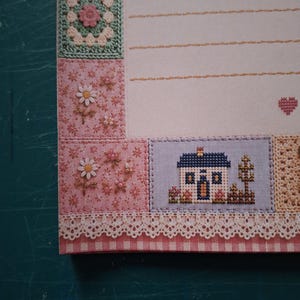 Puede incluir: Una hoja de papel de carta con un borde decorativo de cuadrados de ganchillo, motivos florales y una casa en punto de cruz. El papel incluye l&iacute;neas horizontales discontinuas para escribir y un peque&ntilde;o icono de coraz&oacute;n rojo a la derecha.