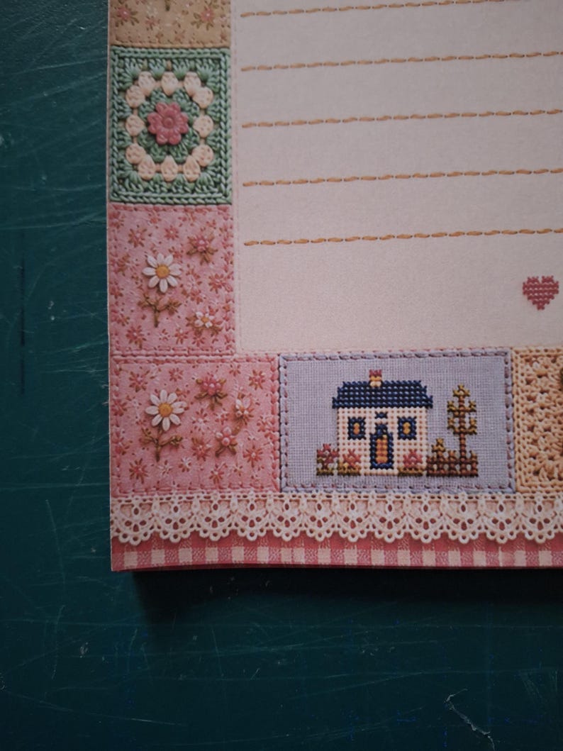 Puede incluir: Una hoja de papel de carta con un borde decorativo de cuadrados de ganchillo, motivos florales y una casa en punto de cruz. El papel incluye l&iacute;neas horizontales discontinuas para escribir y un peque&ntilde;o icono de coraz&oacute;n rojo a la derecha.
