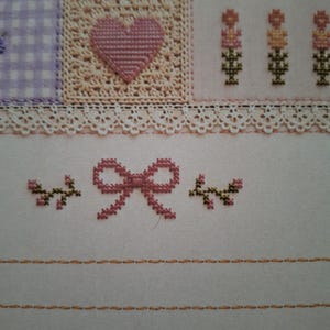 Puede incluir: Una pieza de tela con bordado de punto de cruz. La secci&oacute;n superior muestra un cuadrado de cuadros morados, un cuadrado beige con un coraz&oacute;n rosa y motivos florales. Debajo, un borde de encaje blanco sobre un lazo rosa y peque&ntilde;as ramas.