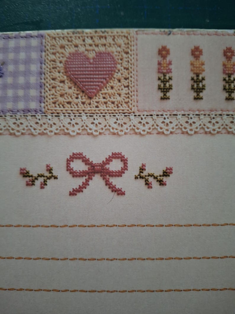 Puede incluir: Una pieza de tela con bordado de punto de cruz. La secci&oacute;n superior muestra un cuadrado de cuadros morados, un cuadrado beige con un coraz&oacute;n rosa y motivos florales. Debajo, un borde de encaje blanco sobre un lazo rosa y peque&ntilde;as ramas.