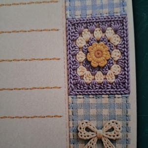 Puede incluir: Una hoja de papel de carta con una zona de escritura beige claro y l&iacute;neas horizontales cosidas en hilo dorado. El borde derecho muestra una franja vertical de tela de cuadros vichy azul y blanca, un cuadrado de ganchillo morado con una flor amarilla y un lazo crema.