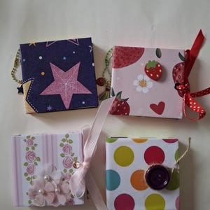 Puede incluir: Cuatro mini cuadernos cuadrados con cubiertas distintas. Uno presenta un patrón de estrellas azul oscuro, otro un estampado de fresas rosa con una cinta roja, uno tiene rosas y flores de tela, y el último muestra lunares coloridos con un botón morado.