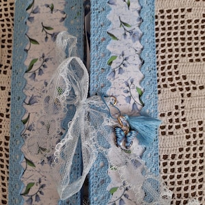 May include: A handmade journal with a light blue crocheted border and a floral patterned interior. The journal is tied with a white lace ribbon and features a butterfly charm and tassel. The overall design is delicate and feminine.