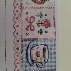 以下が含まれることがあります： 青いギンガムチェックの背景にピンクのハートが描かれた白いティーカップのクロスステッチ模様。その上にはピンクのリボンと小さな青い花が描かれた正方形があり、全体がピンクのスカラップレースの縁取りで囲まれています。