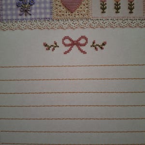 Puede incluir: Papel de carta con un borde superior de tres cuadrados en punto de cruz: un cuadro de vichy morado con flores azules, un cuadro beige con un coraz&oacute;n rosa y un cuadro blanco con tres ramilletes florales. Debajo hay un borde de encaje blanco.