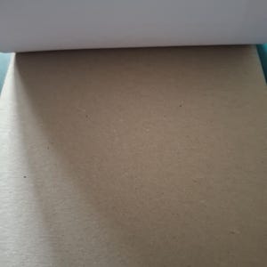 Puede incluir: Un bloc rectangular de papel kraft de color marr&oacute;n claro con una textura lisa y mate. El borde superior muestra una gruesa tira de encuadernaci&oacute;n blanca, mientras que el resto de la superficie presenta un color beige neutro y uniforme.
