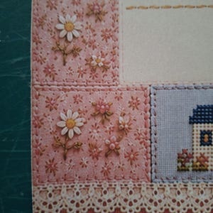 Puede incluir: Una tarjeta decorativa de papel con un dise&ntilde;o de patchwork y patrones florales bordados en rosa, blanco y verde. La tarjeta incluye un peque&ntilde;o motivo de casa en punto de cruz, un &aacute;rea de escritura color crema y un borde de encaje blanco.