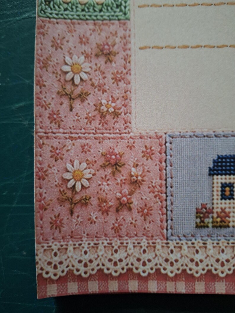 Puede incluir: Una tarjeta decorativa de papel con un dise&ntilde;o de patchwork y patrones florales bordados en rosa, blanco y verde. La tarjeta incluye un peque&ntilde;o motivo de casa en punto de cruz, un &aacute;rea de escritura color crema y un borde de encaje blanco.