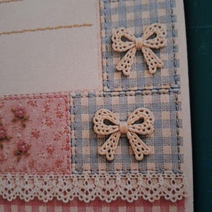 Puede incluir: Una hoja de papel de carta con un dise&ntilde;o de patchwork que incluye cuadros vichy azul claro, una secci&oacute;n de flores rosas y un borde de encaje color crema. Dos lazos decorativos color crema est&aacute;n fijados sobre los cuadros y el papel tiene l&iacute;neas.
