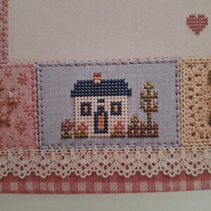 May include: A cross-stitch embroidery piece featuring a small house with a dark blue roof, white walls, and yellow windows. The house is set against a light blue background with a brown fence and tree, bordered by pink floral fabric and cream lace.
