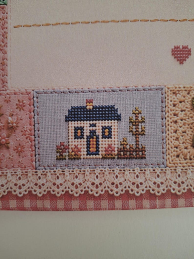 May include: A cross-stitch embroidery piece featuring a small house with a dark blue roof, white walls, and yellow windows. The house is set against a light blue background with a brown fence and tree, bordered by pink floral fabric and cream lace.
