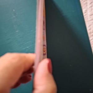 Puede incluir: Un folleto rectangular delgado con una cubierta de color verde azulado oscuro y un lomo claro. El perfil lateral revela varias p&aacute;ginas internas con peque&ntilde;as ilustraciones cuadradas de colores visibles en el borde del papel.
