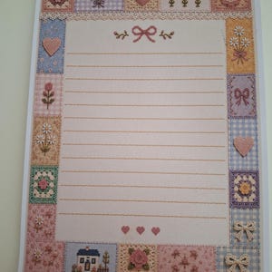 Puede incluir: Una hoja de papel de carta rectangular con un &aacute;rea central de l&iacute;neas horizontales para escribir, enmarcada por un borde de varios cuadrados. Los cuadrados muestran patrones de punto de cruz de flores, corazones, lazos y una casa en tonos pastel.
