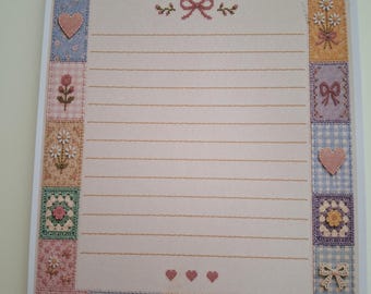 May include: A rectangular sheet of stationery featuring a central area with horizontal lines for writing, framed by a border of various square patches. The patches display cross-stitch patterns of flowers, hearts, bows, and a house in pastel colours.